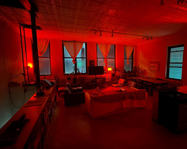 The room is illuminated with red lighting, featuring furniture arranged around a central table and windows covered by sheer curtains.