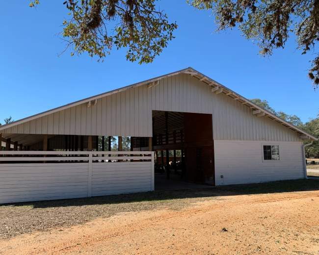 A large white barn with a peaked roof sits beside a dirt path under clear blue skies.