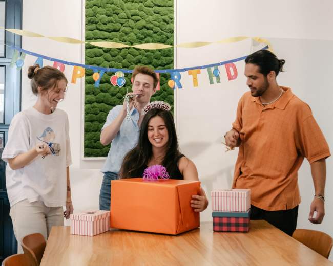 A group of four people gathers around a table in a bright room, celebrating a birthday as one person opens a large orange gift box.