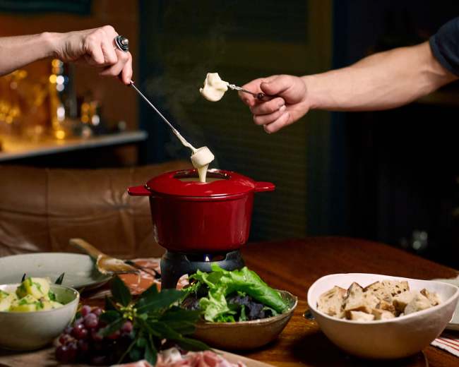 Two people are using fondue forks to dip pieces of cheese into a red fondue pot, surrounded by various ingredients on a wooden table.