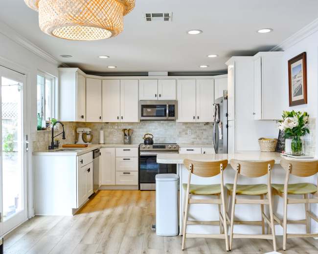 The image shows a modern kitchen with white cabinetry, stainless steel appliances, and a breakfast bar with three wooden stools.