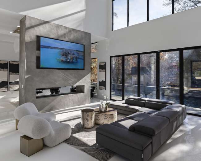The image shows a modern living room with large windows, a dark sectional sofa, a round coffee table, and an entertainment system mounted on the wall.
