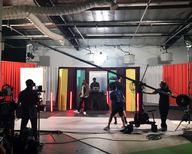A film crew sets up equipment on a colorful studio set with a central stage for a live performance.