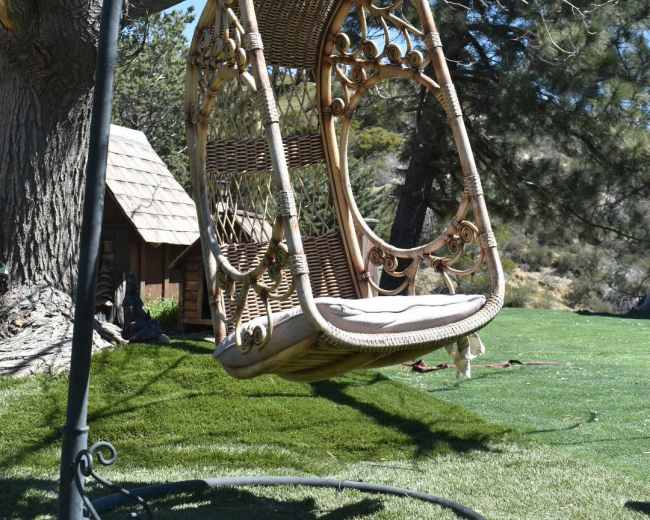 A wicker swing hangs from a metal frame near a bare tree in a grassy area.