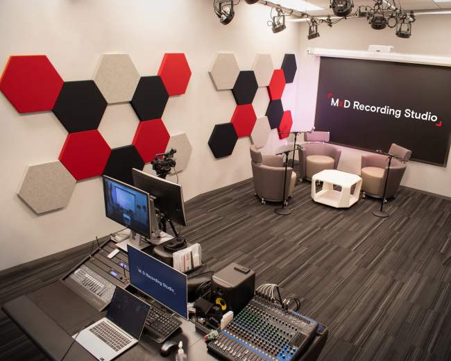 The image shows a modern recording studio with hexagonal sound panels on the walls, two armchairs and a coffee table in the center, and a control area with multiple monitors and audio equipment.