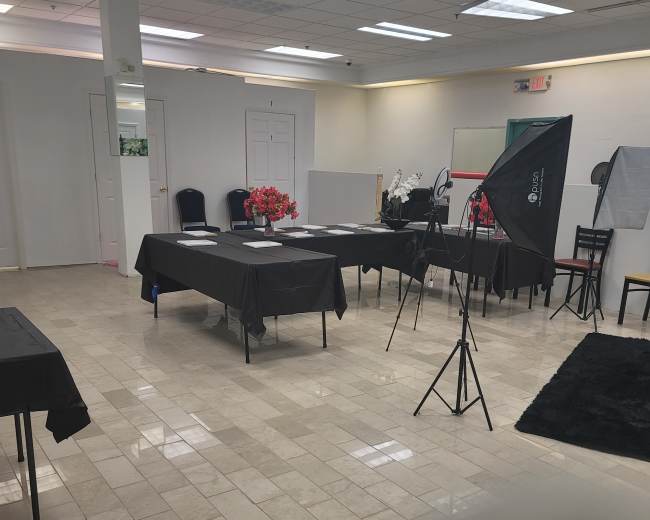 A spacious room with several tables covered in black cloth, equipped with flowers, and a lighting setup on a tripod.