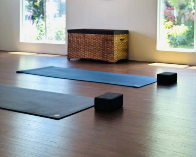 The image shows a yoga studio with two mats, two yoga blocks, a round mirror, and a wicker storage box, illuminated by natural light from large windows.