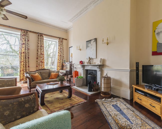 The image shows a living room with a fireplace, seating area, and a large window overlooking a tree-filled landscape.