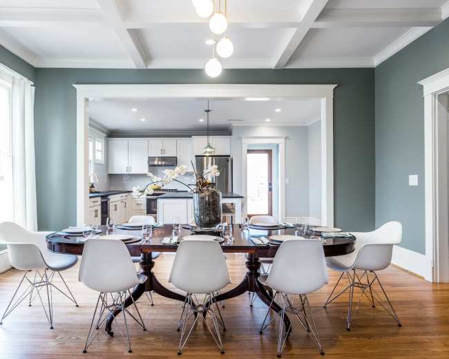 A modern dining room features a large wooden table surrounded by white chairs, with a kitchen visible in the background.