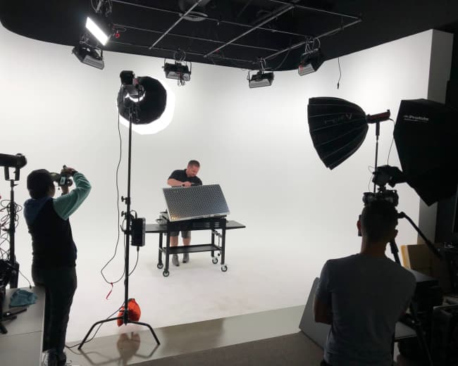 A person is setting up a project on a table in a well-lit studio surrounded by cameras and lighting equipment.