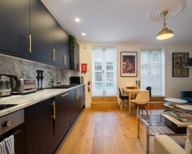 The image shows a modern kitchen and living area featuring dark cabinetry, a marble countertop, and a small dining table beside large windows with white blinds.