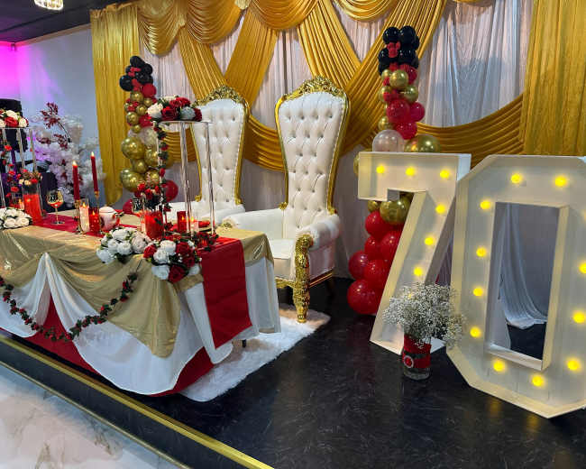 The image shows a decorative event setup featuring a gold and white throne-like chair, a table adorned with red and white flowers, and a lighted centerpiece displaying the number 70, surrounded by balloons.