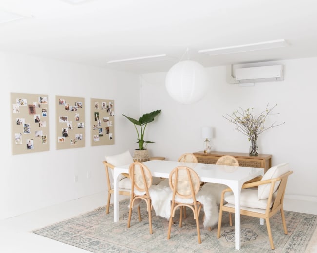A bright, minimally decorated dining area features a long table surrounded by chairs, with photos on the wall and a large mirror reflecting the space.