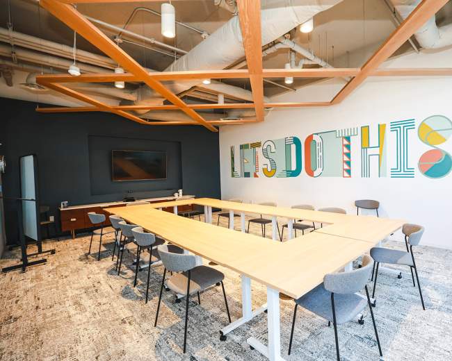 The image shows a modern conference room with a long table, multiple chairs, and a wall featuring colorful text that reads "LET'S DO THIS."