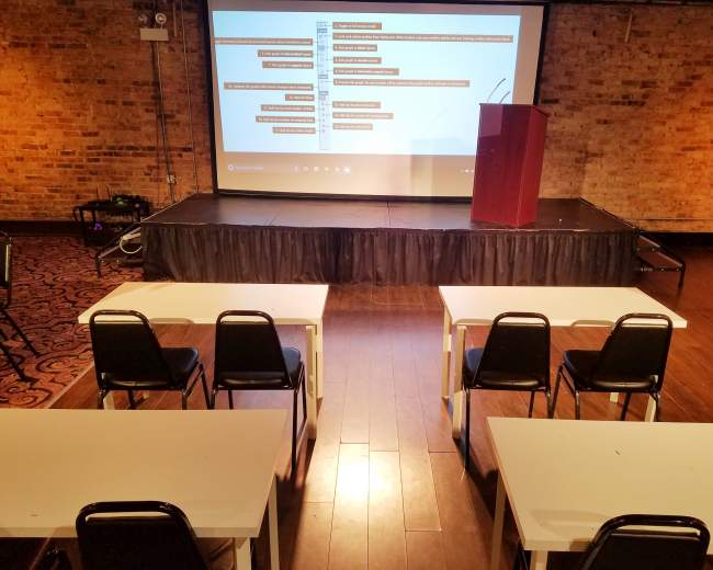 The image shows a presentation setup in a room with a stage, a podium, and several rows of empty tables and chairs facing a screen displaying content.