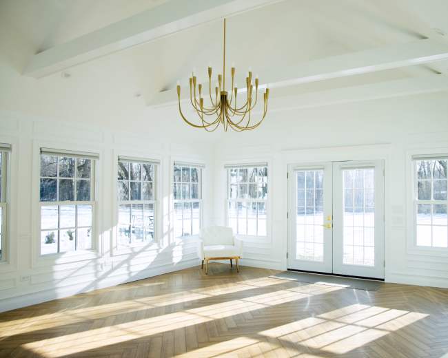 The image shows a bright, empty room with large windows, a chandelier, and a herringbone wood floor.
