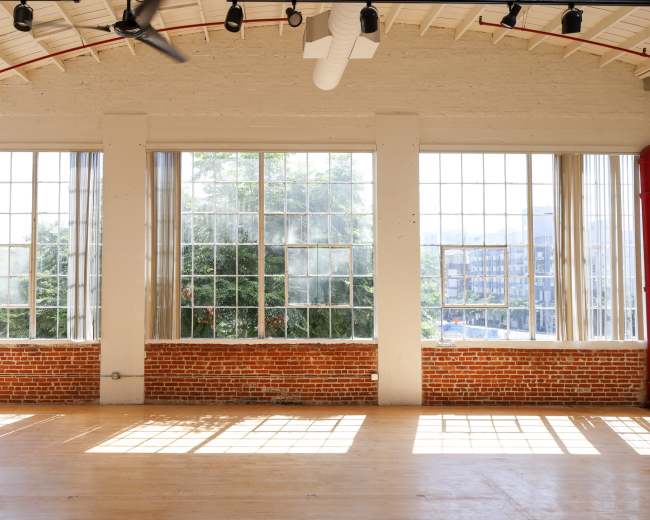 The image shows a spacious room with large windows that allow natural light to illuminate the wooden floor and reveal greenery outside.