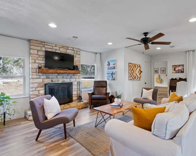 The image shows a cozy living room with a stone fireplace, comfortable seating arrangements, and large windows allowing natural light to enter.