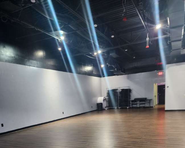 The image shows an empty room with a wooden floor, black walls, and multiple overhead lights illuminating the space.