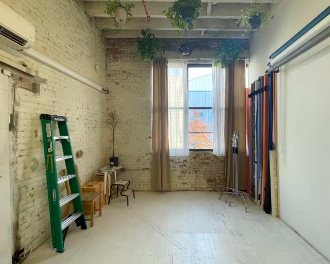 A spacious, empty room with exposed brick walls, a large window, and a ladder, along with a few chairs and draped fabrics.