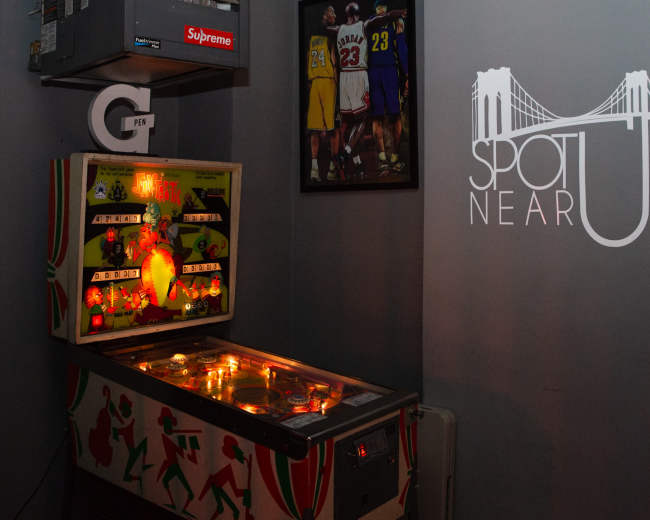 A vintage pinball machine is illuminated in a dimly lit corner, next to a wall featuring a framed image of basketball players and a logo with the text "SPOT NEAR U."