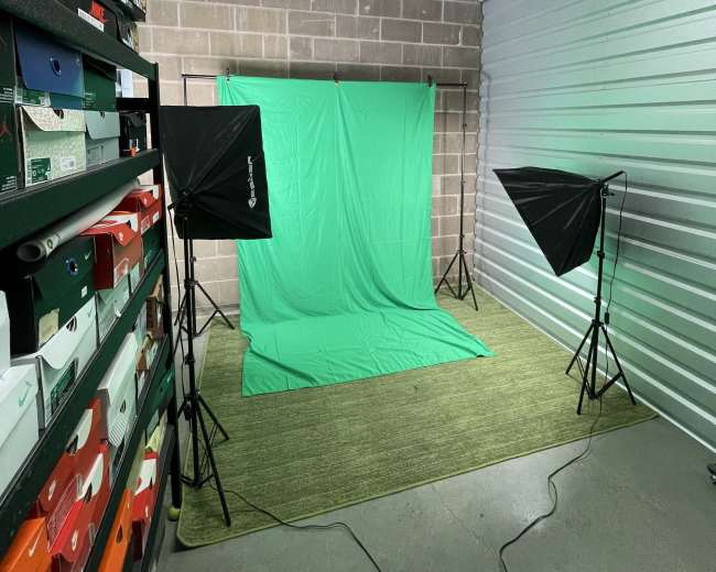 A storage unit contains a green backdrop set up for photography, flanked by two softbox lights and surrounded by stacked shoeboxes.