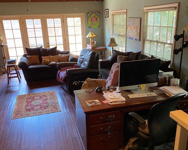 The image shows a living room with a dark couch, a computer desk, and a large window letting in natural light.