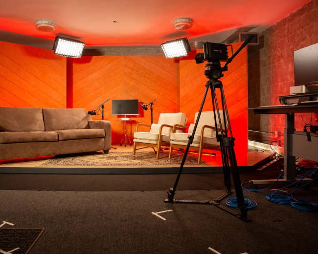 The image shows a studio setup with a sofa, two armchairs, a coffee table, and lighting equipment against an orange backdrop.