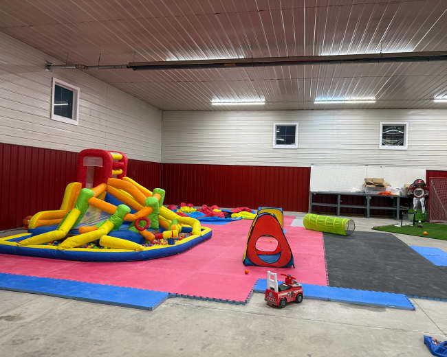The image shows an indoor play area featuring colorful inflatable structures, soft play mats, and various children's toys scattered across the floor.