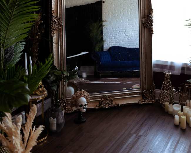A large ornate mirror reflects a blue couch in a dimly lit room decorated with plants and candles.