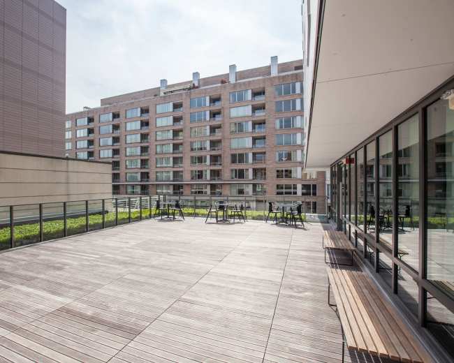 The image shows a spacious wooden terrace with metal railings, surrounded by modern buildings featuring large windows.