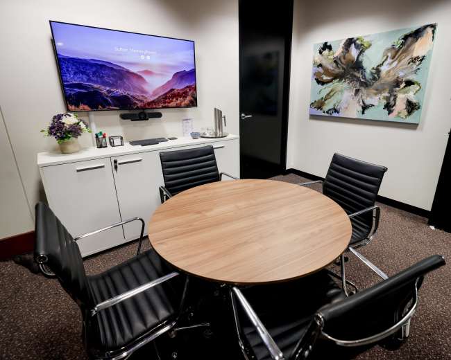 The image shows a small meeting room with a round wooden table, four black leather chairs, a wall-mounted television, and abstract art on the wall.