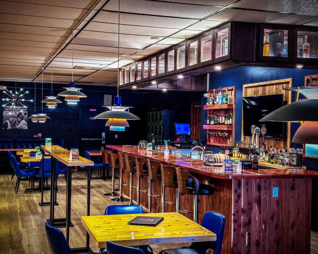 The image shows a modern bar interior with wooden tables, high stools, and a well-stocked bar counter illuminated by pendant lights.