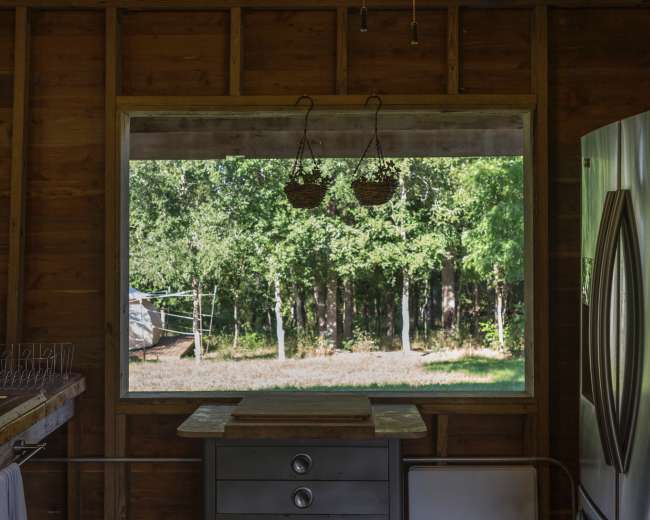 A rustic kitchen with a wooden interior, featuring a metal table, a refrigerator, and a large window overlooking a wooded area.