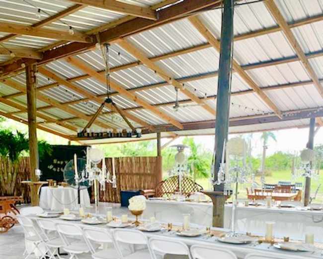 The image shows a banquet setting with several tables covered in white cloth and arranged with plates and cutlery, all under a wooden structure.