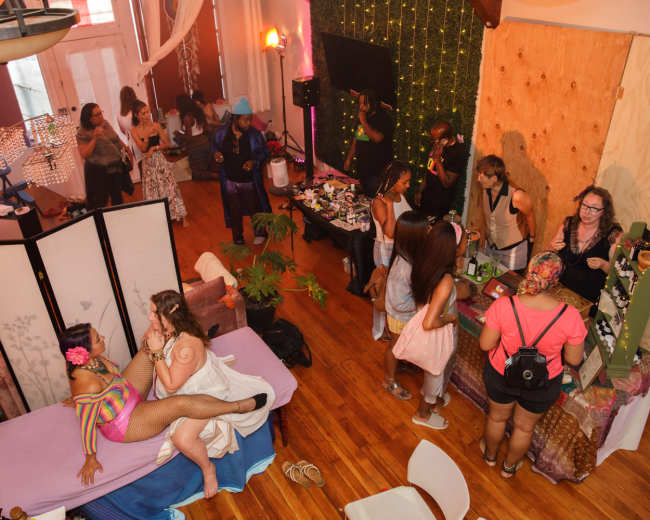 A lively indoor event space with diverse attendees engaged in various activities, including a performance area and a vendor setup.