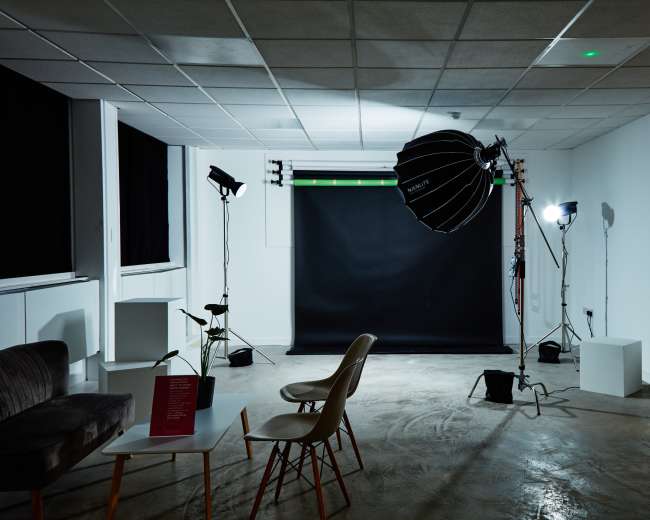 The image shows a photography studio with a black backdrop, lighting equipment, and a few pieces of furniture including a sofa and chairs.