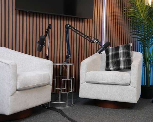 The image features two modern, light gray swivel chairs positioned next to a small table, with a plant and lighted backdrop in a studio setting.