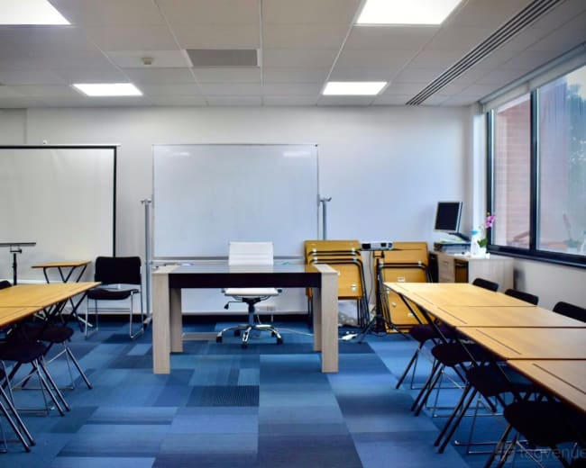 A modern meeting room features a central table with a chair, surrounded by several tables arranged in a U-shape, and equipped with a whiteboard and storage cabinet.