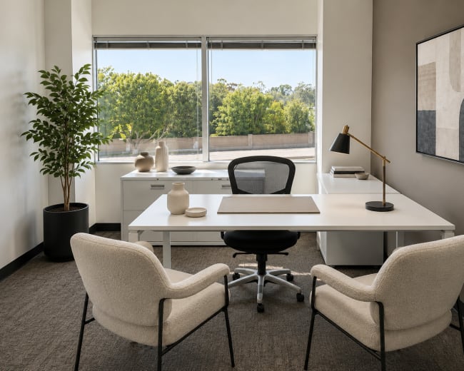 The image shows a modern office space with a white desk and black rolling chair, flanked by two light-colored armchairs, a potted plant, and a large window overlooking greenery.