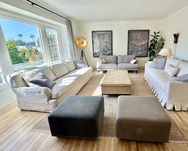 A spacious living room features three sofas, a central coffee table, and large windows allowing natural light to illuminate the wooden flooring.