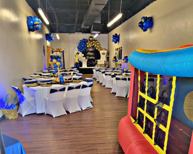 The image shows a decorated event space with tables covered in blue and gold, a bounce house in the corner, and balloon arrangements on the walls.