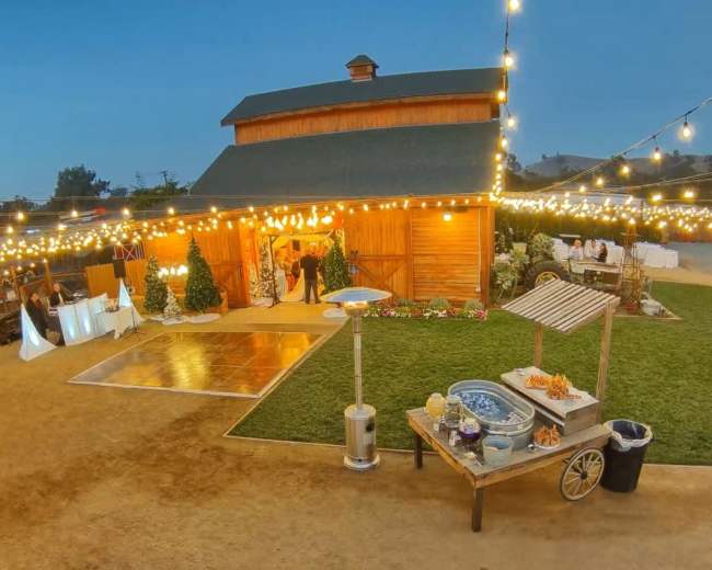 A rustic barn with string lights illuminates an outdoor area featuring a dance floor, tables, and a food station.