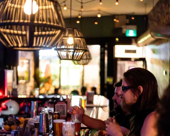 Two patrons sit at a bar, enjoying drinks and snacks under warm pendant lighting.