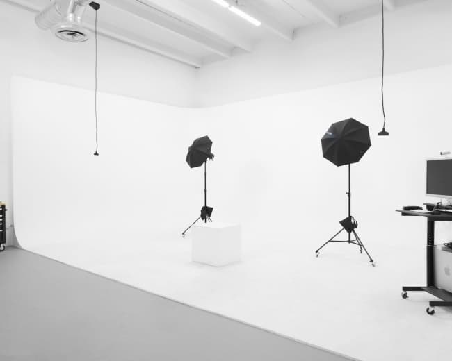 The image shows a minimalist photography studio with two black umbrella lights on tripods facing a white backdrop and a small pedestal in the center.