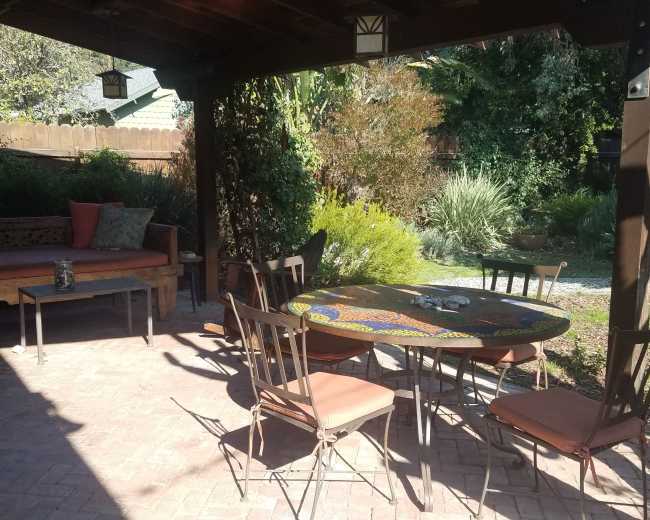 The image shows a shaded outdoor patio area with a round table, six chairs, and a wooden bench surrounded by greenery.