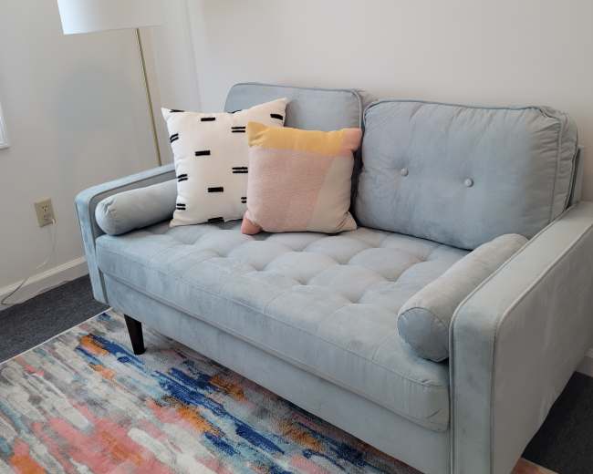 A light blue sofa with decorative pillows sits on a colorful rug next to a tall lamp in a bright room.