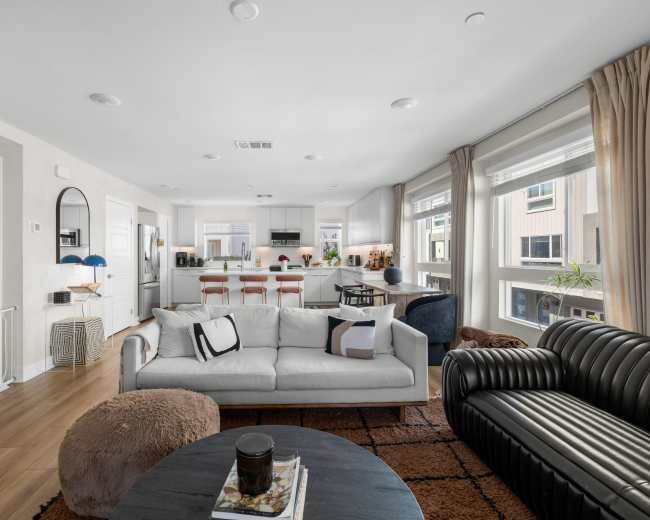 The image shows a modern living room area connected to a kitchen, featuring a gray couch, a black leather sofa, and a circular coffee table.