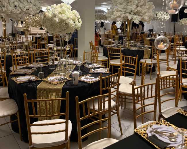 The image shows an elegantly decorated banquet hall with round tables draped in black and gold, adorned with floral centerpieces and crystal chandeliers overhead.