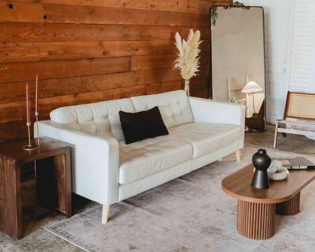 The image shows a modern living room with a white leather sofa, a wooden wall, a large mirror, a small table with decorative items, and a rug on a concrete floor.